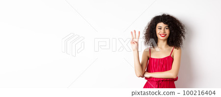 Beautiful lady with curly hair, wearing elegant red dress and showing number three, making order, standing over white background 100216404
