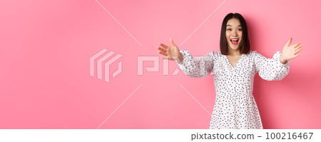 Happy asian girl stretching hands to camera, reaching for hug, smiling and wanting to hold something, standing over pink background in dress 100216467
