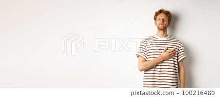 Young bearded redhead man holding hand on heart and looking patriotic, standing during national anthem, standing over white background 100216480