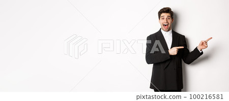 Image of attractive smiling guy dressed for new year party, pointing fingers right and showing advertisement, looking impressed standing over white background Image of attractive smiling guy dressed for new year party, pointing fingers right and showing advertisement, looking impressed standing over white background 100216581