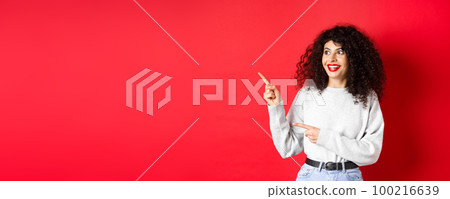 Portrait of excited curly woman with red lips, looking and pointing left at logo with amazed face, checking out promo deal, standing on studio background 100216639