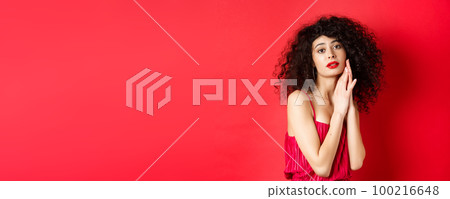 Romantic sensual woman with curly hair, wearing evening dress, posing seductive on red background 100216648