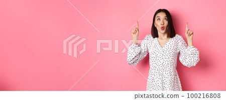 Cute korean girl in beautiful dress saying wow, looking and pointing fingers up, intrigued in promo offer, standing over pink background 100216808