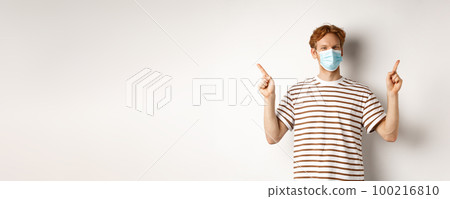 Covid, virus and social distancing concept. Handsome young man with red hair, wear face mask and pointing sideways at two promo offers, white background 100216810
