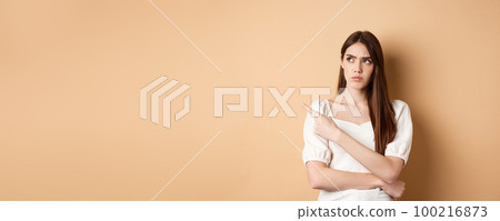 Suspicious woman in dress looking, pointing left, frowning and staring with disbelief and doubts, standing on beige background 100216873