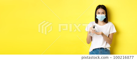 Covid, health care and pandemic concept. Cute asian woman in medical mask using hand sanitizer to disinfect palm, standing over yellow background Covid, health care and pandemic concept. Cute asian woman in medical mask using hand sanitizer to disinfect palm, standing over yellow background 100216877