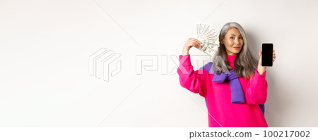 Fashionable asian senior woman showing money dollars and blank smartphone screen, demonstrate online shop, standing over white background Fashionable asian senior woman showing money dollars and blank smartphone screen, demonstrate online shop, standing over white background 100217002