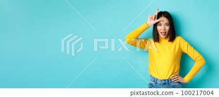 Asian girl winner mocking over lost team, showing loser sign on forehead and sticking tongue ignorant, standing over blue background Asian girl winner mocking over lost team, showing loser sign on forehead and sticking tongue ignorant, standing over blue background 100217004