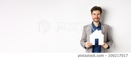 Real estate and insurance concept. Salesman in grey suit showing paper house cutout, selling property, smiling friendly at camera, white background Real estate and insurance concept. Salesman in grey suit showing paper house cutout, selling property, smiling friendly at camera, white background 100217017