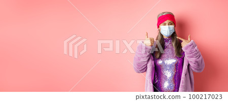 Covid-19, social distancing and fashion concept. Smiling asian senior woman in fashionable clothes and respirator, pointing at face mask and show thumb-up, pink background 100217023