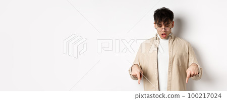 Amazed young guy in glasses look and point down, open mouth and say wow impressed, checking out discount offer, standing on white background 100217024