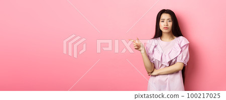 Skeptical chinese girl pointing finger left, looking unamused and serious, showing offer, standing in dress on pink background 100217025