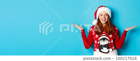Happy holidays and Christmas concept. Cheerful redhead girl in Santa hat and xmas sweater, raising hands on copy spaces, holding something on blue background 100217028