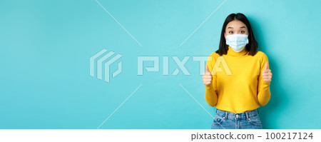 Covid-19, social distancing and pandemic concept. Excited asian woman showing thumbs up and looking impressed, praising good deal, wearing face mask, blue background 100217124