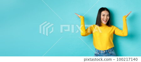 Beauty and fashion concept. Stylish beautiful asian girl demonstrate promotion, raising hands up and smiling, showing something on blue background Beauty and fashion concept. Stylish beautiful asian girl demonstrate promotion, raising hands up and smiling, showing something on blue background 100217140