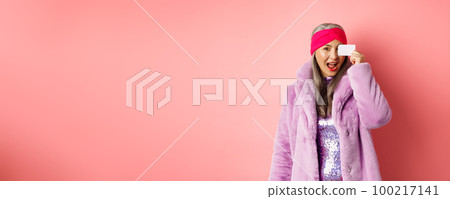 Shopping and fashion concept. Beautiful asian middle-aged woman showing plastic credit card on face and looking excited at camera, standing over pink background 100217141