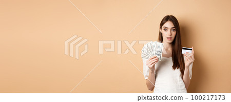 Excited woman holding dollar bills and plastic credit card, looking amazed at camera, standing on beige background 100217173