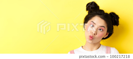Close up portrait of funny asian teen girl with glamour makeup and hairstyle, squinting eyes and pucker lips silly, standing on yellow background Close up portrait of funny asian teen girl with glamour makeup and hairstyle, squinting eyes and pucker lips silly, standing on yellow background 100217218