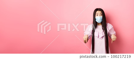 Healthy people and covid-19 pandemic concept. Excited asian girl in face mask pointing fingers down, showing advertisement with interest, standing against pink background 100217219