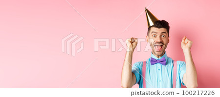 Holidays and celebration concept. Cheerful young man celebrating birthday party in cone hat and bow-tie, shouting yes with excitement and making fist pump gesture, pink background Holidays and celebration concept. Cheerful young man celebrating birthday party in cone hat and bow-tie, shouting yes with excitement and making fist pump gesture, pink background 100217226