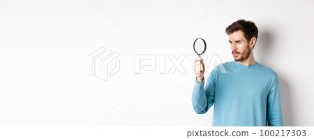 Young handsome man look through magnifying glass with curious face, investigating or searching for something, standing on white background Young handsome man look through magnifying glass with curious face, investigating or searching for something, standing on white background 100217303