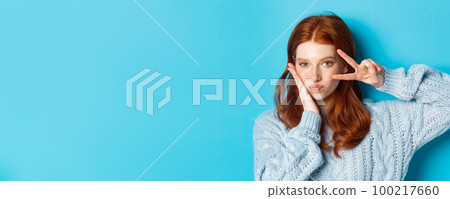 Close-up of beautiful and sassy girl with red hair, showing peace kawaii sign and gazing at camera, standing over blue background Close-up of beautiful and sassy girl with red hair, showing peace kawaii sign and gazing at camera, standing over blue background 100217660