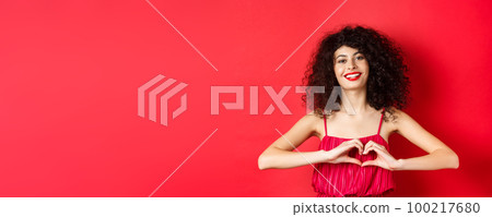Lovers day. Beautiful woman celebrating valentines, showing heart sign and smiling, standing in romantic red dress on studio background Lovers day. Beautiful woman celebrating valentines, showing heart sign and smiling, standing in romantic red dress on studio background 100217680