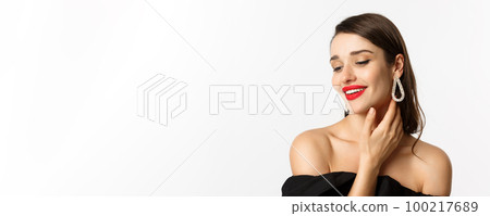 Fashion and beauty concept. Close-up of tender woman in black dress and earrings, gently touching face and smiling, looking down coquettish, standing over white background 100217689