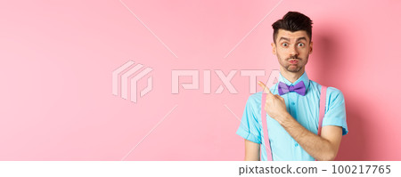 Impressed young man showing awesome promo offer, raising eyebrows and look interested, pointing fingers at upper left corner, pink background 100217765