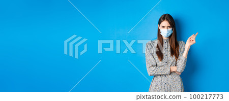 Covid-19, social distancing and healthcare concept. Angry young woman in medical mask frowning, pointing right at bad thing, condemn non-maskers, blue background 100217773