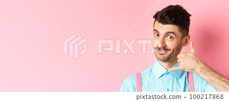 Close-up of handsome young guy with moustache, showing phone gesture, asking to call him, standing on pink background Close-up of handsome young guy with moustache, showing phone gesture, asking to call him, standing on pink background 100217868