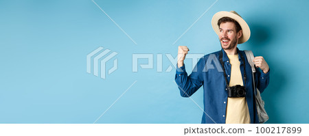 Cheerful tourist rejoicing on vacation, celebrating summer holidays, showing fist pump gesture and saying yes with satisfied face, looking aside, blue background 100217899