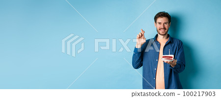 Celebration and holiday concept. Hopeful young man making wish on birthday cake, cross fingers for good luck and smiling positive, standing on blue background Celebration and holiday concept. Hopeful young man making wish on birthday cake, cross fingers for good luck and smiling positive, standing on blue background 100217903