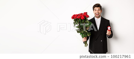 Handsome smiling man in black suit, holding roses and engagement ring, making a proposal to marry him, standing against white background Handsome smiling man in black suit, holding roses and engagement ring, making a proposal to marry him, standing against white background 100217961