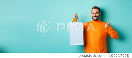 Handsome smiling man showing thumbs-up and shopping bag, recommending store, standing over blue background Handsome smiling man showing thumbs-up and shopping bag, recommending store, standing over blue background 100217968