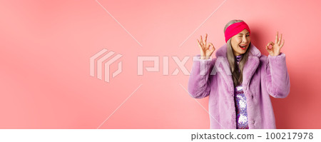 Fashion and shopping concept. Stylish asian granny saying all okay and winking. Trendy senior woman in winter fake-fur coat show OK sign, praising good product, pink background 100217978
