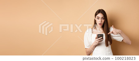 Online shopping concept. Excited girl pointing at smartphone screen and looking at camera amazed, standing against beige background 100218023