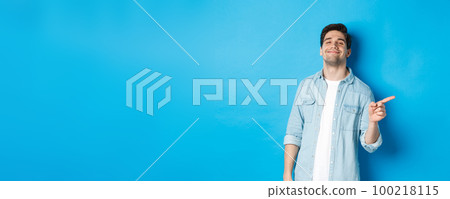 Confident handsome guy smiling sassy, pointing finger right at copy space, showing advertisement, standing over blue background 100218115