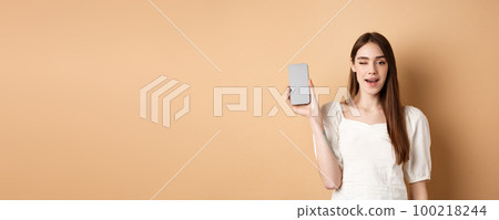Cheerful stylish girl showing smartphone app, demonstrate empty phone screen and winking, standing on beige background 100218244