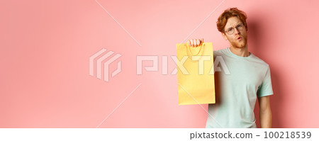 Handsome young man in glasses looking sassy at camera, showing yellow shopping bag, telling about discounts in shop, standing over pink background 100218539