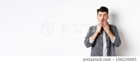 Shocked caucasian guy gasping alarmed, cover mouth with hands and staring at something terrible, witness bad accident, say oh my god, standing on white background 100220065