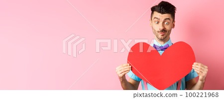 Valentines day concept. Cute boyfriend looking at big red heart, waiting for lover on romantic date, pink background 100221968