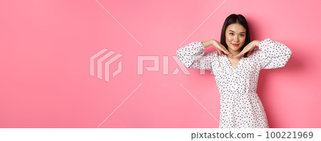 Beautiful asian woman in trendy dress making cute face, holding hands near jawline and gazing coquettish at camera, standing over pink background 100221969