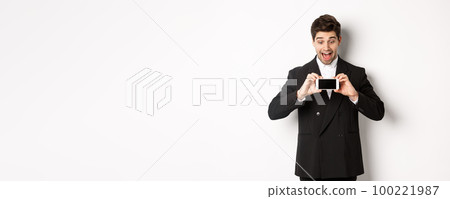 Portrait of excited handsome man showing smartphone screen, looking amazed at display with advertisement, standing over white background in black suit 100221987