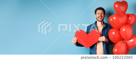 Heartbroken man crying of breakup of valentines day, holding red heart cutout and standing near romantic balloons over blue background Heartbroken man crying of breakup of valentines day, holding red heart cutout and standing near romantic balloons over blue background 100222005