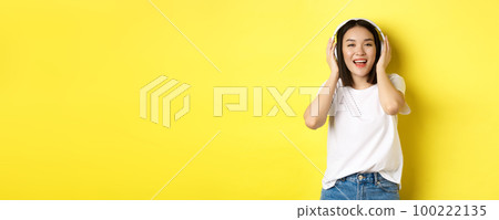 Attractive asian woman enjoying listening music in wireless headphones, smiling pleased and singing along, yellow background 100222135