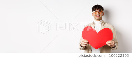 Attractive modern man smiling and looking at camera hopeful, holding big red Valentines heart, waiting for soulmate on lover day date, standing over white background Attractive modern man smiling and looking at camera hopeful, holding big red Valentines heart, waiting for soulmate on lover day date, standing over white background 100222303