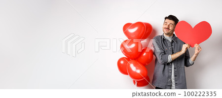 Romantic guy celebrating Valentines day, hugging big red heart card from lover and smiling happy, being in love, standing on white background near red balloons Romantic guy celebrating Valentines day, hugging big red heart card from lover and smiling happy, being in love, standing on white background near red balloons 100222335