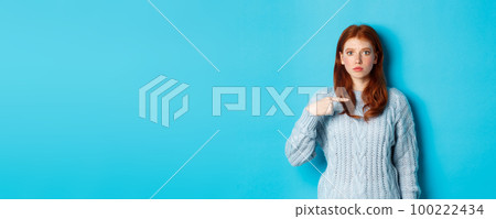Nervous and confused redhead girl pointing at herself, standing in sweater against blue background 100222434