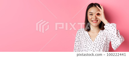 Beauty and lifestyle concept. Close-up of cute smiling asian girl showing okay sign on eye, standing over pink background in dress 100222441